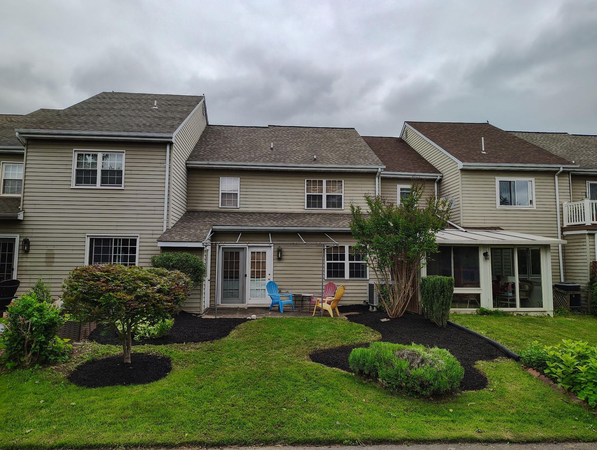 Tan townhouses with siding, gray shingled roofs, and small front lawns with mulch beds and a few bushes under cloudy skies.