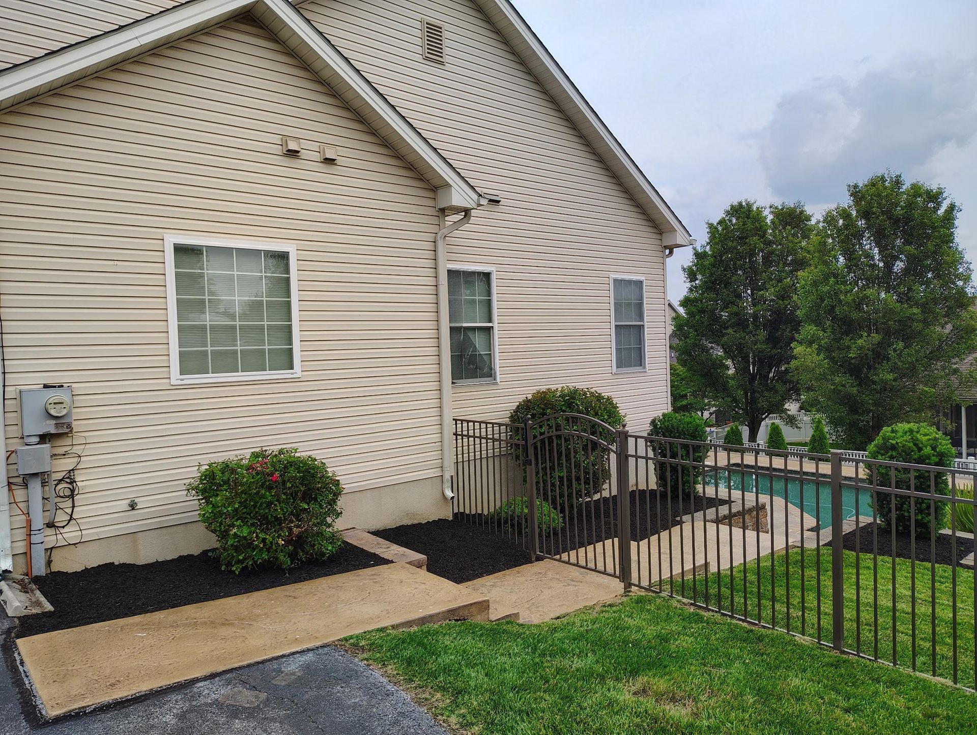 Side view of a beige house with siding, manicured shrubs, a concrete path, and a metal fence bordering a backyard pool.