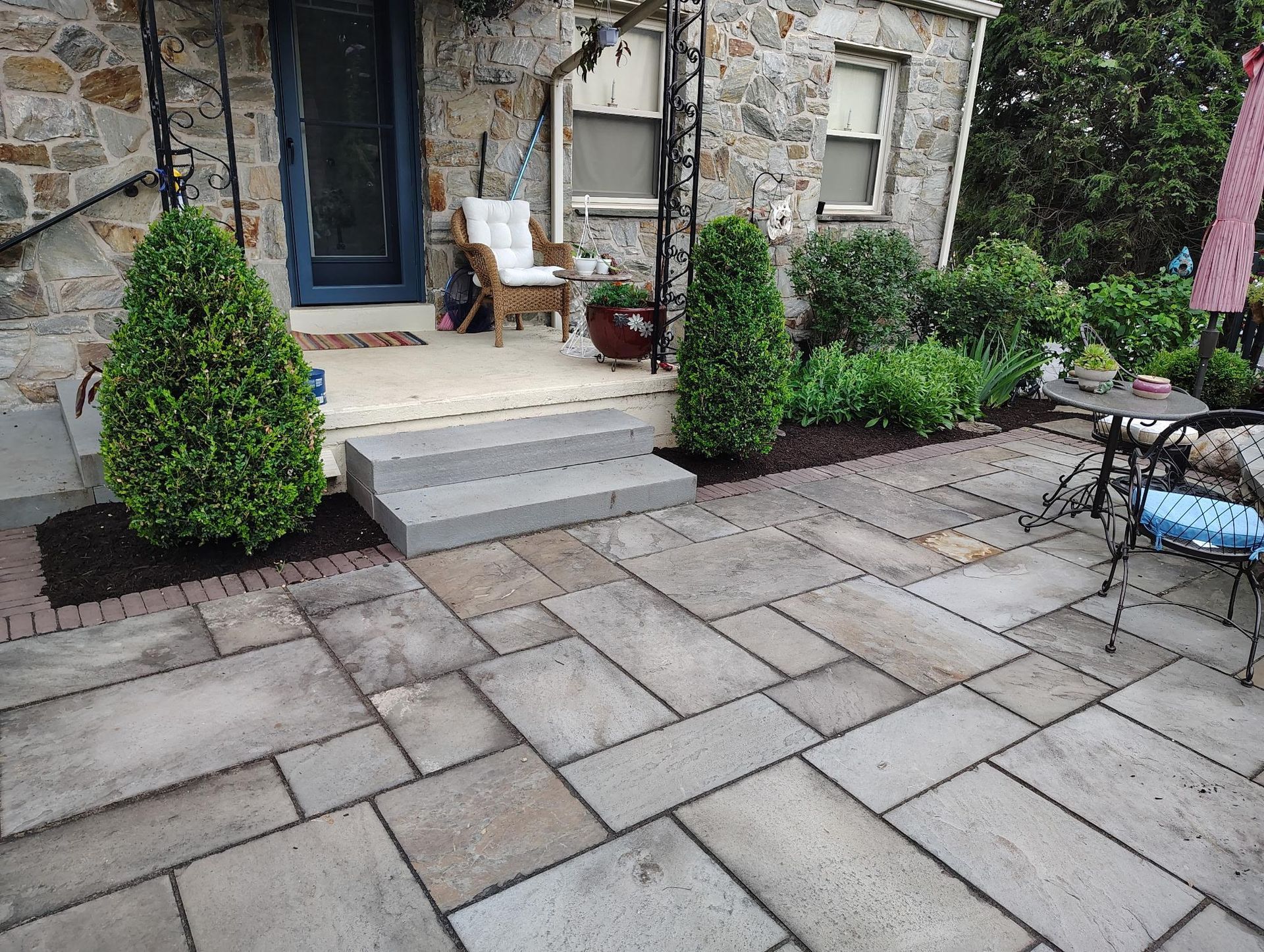 A stone patio with a light gray flagstone floor leads to a small porch and home entrance, flanked by two green shrubs.