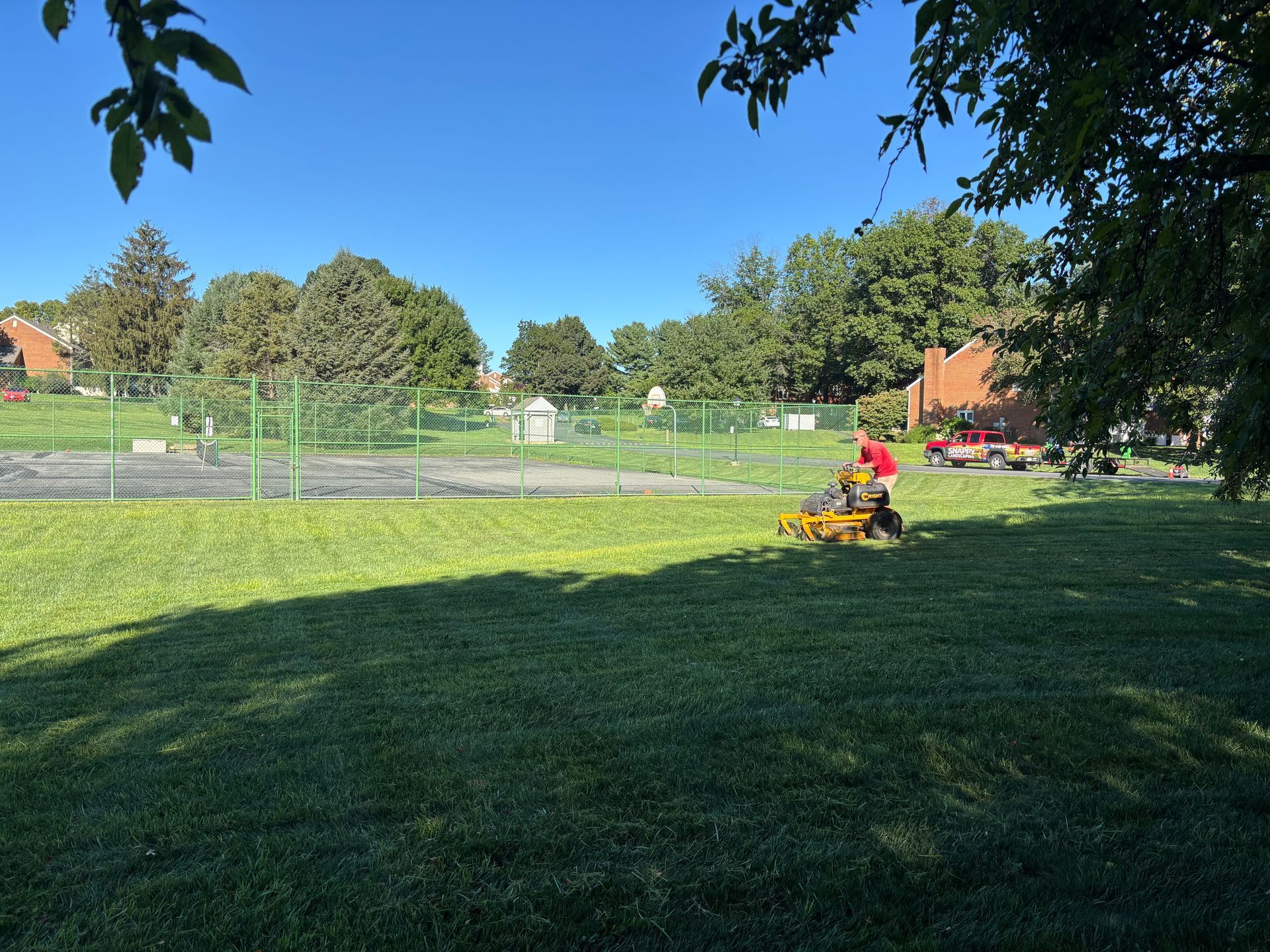 Person mowing a grassy field near a fenced playground on a sunny day