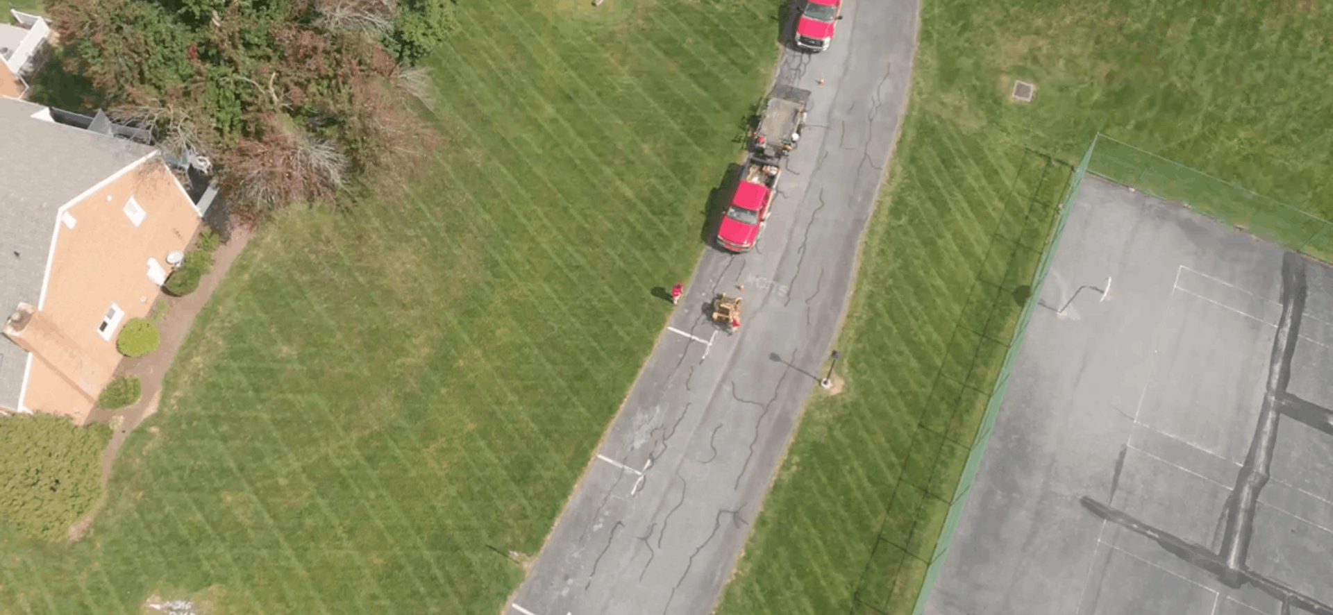 Aerial view of a red car and a few people on a paved road beside a grassy lawn and a parking lot.