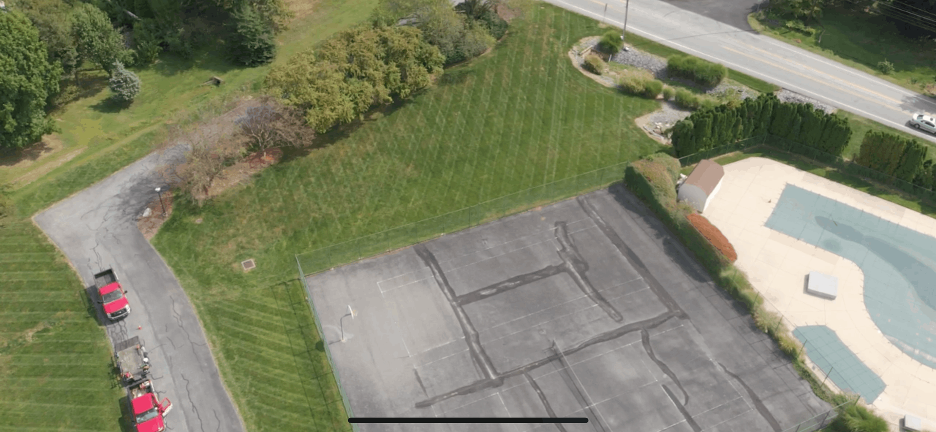 Aerial view of a paved sports court next to a swimming pool and grassy area, bordered by a road and trees.