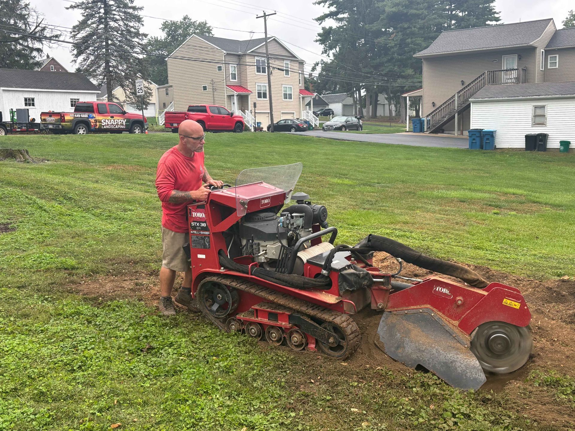 Person operating a red tracked stump grinder on a grassy lot near houses.
