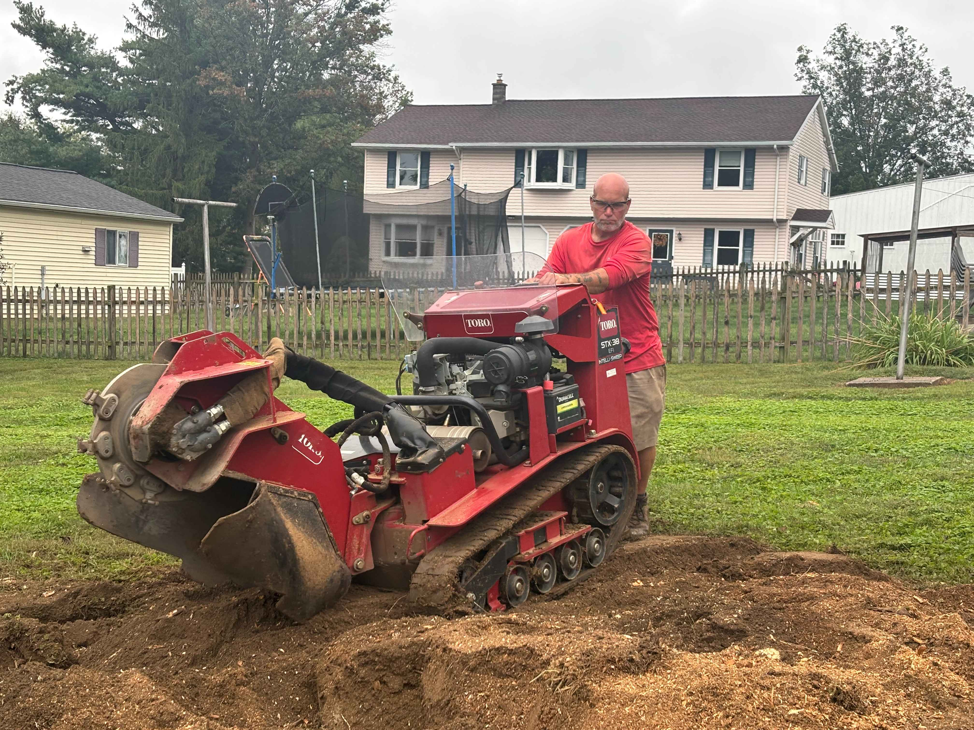 Person in red operating a red skid-steer loader on muddy ground near a house and fence.
