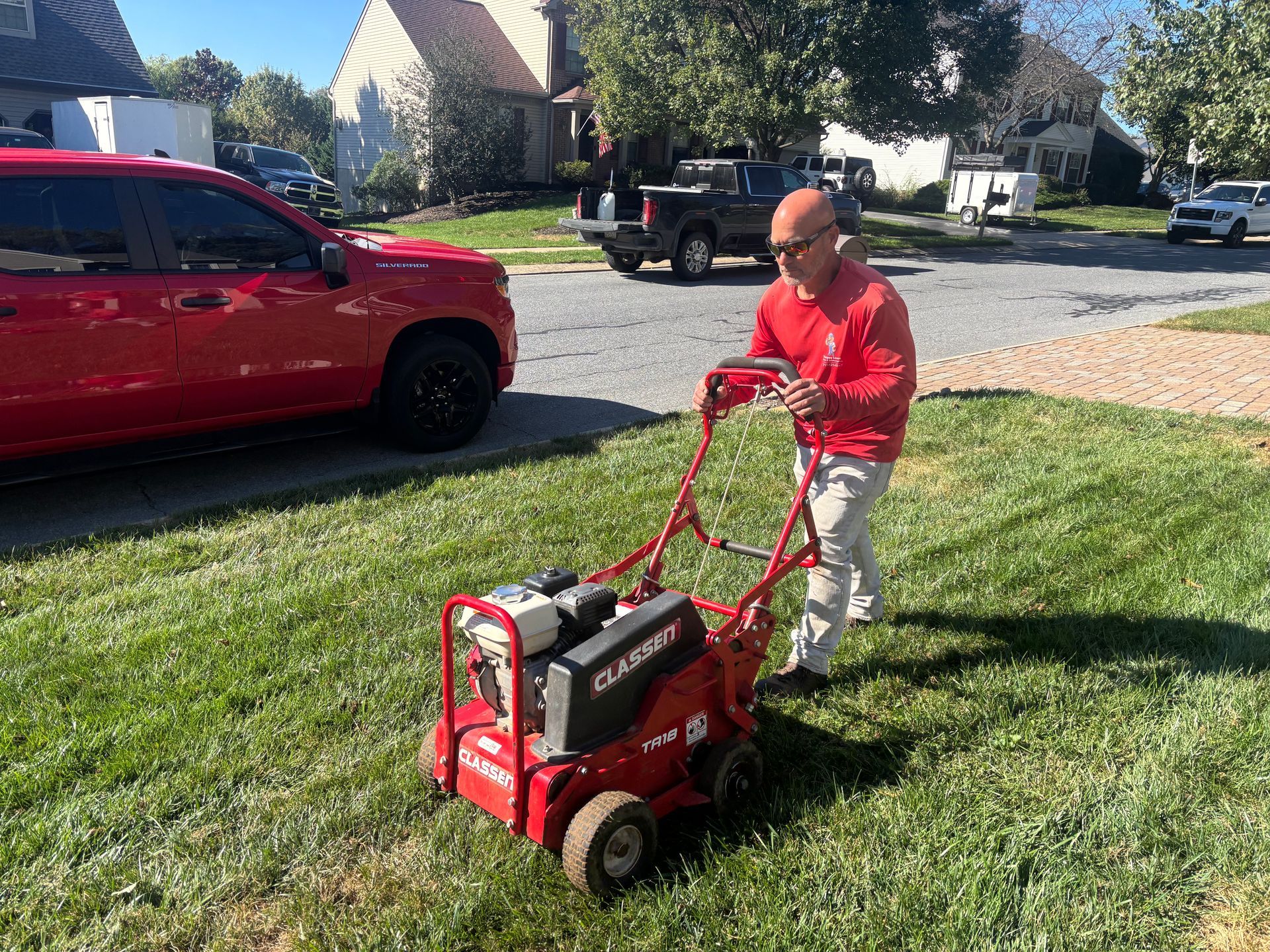Man in a red shirt pushing a red lawn machine across a grassy yard beside a red truck.