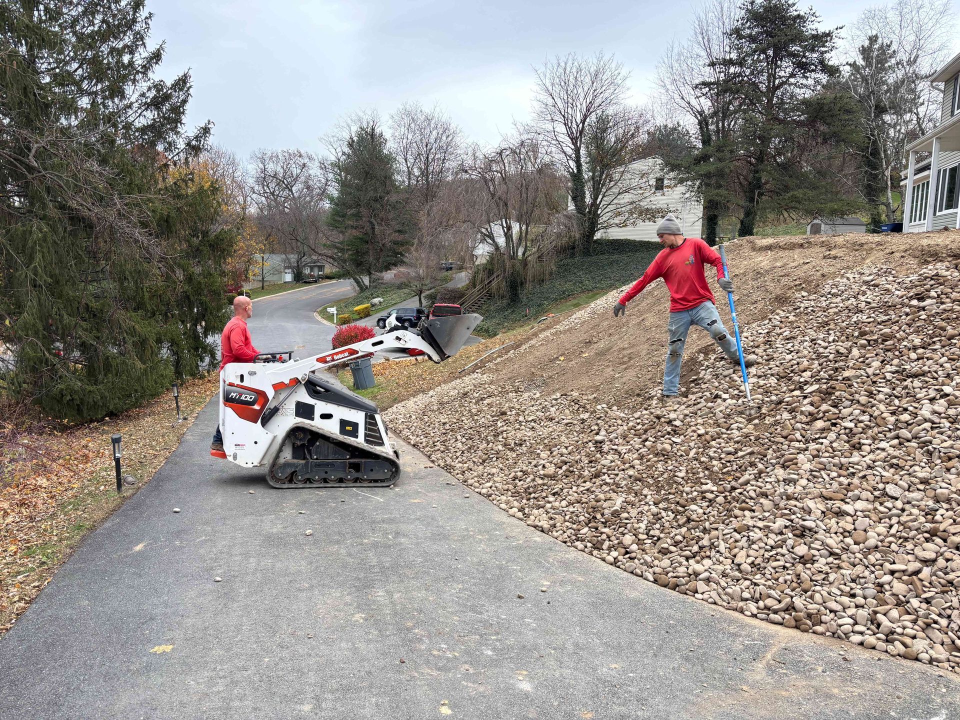 Two workers in red shirts work on a landscaped hill with a small track loader, one operating the machine and one on foot.