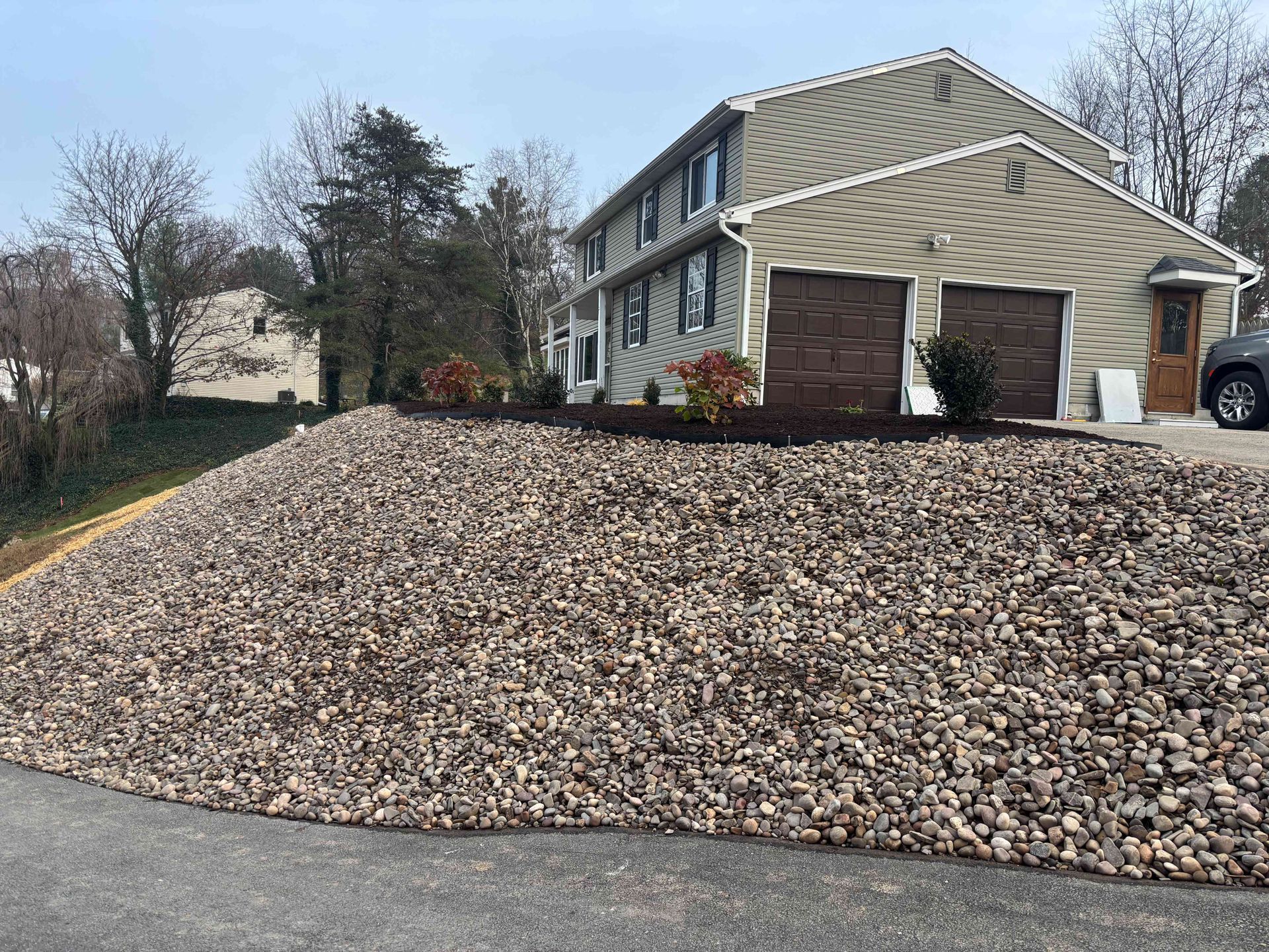 A tan, two-story house with two brown garage doors sits atop a steep, sloped front yard covered in large landscaping rocks.