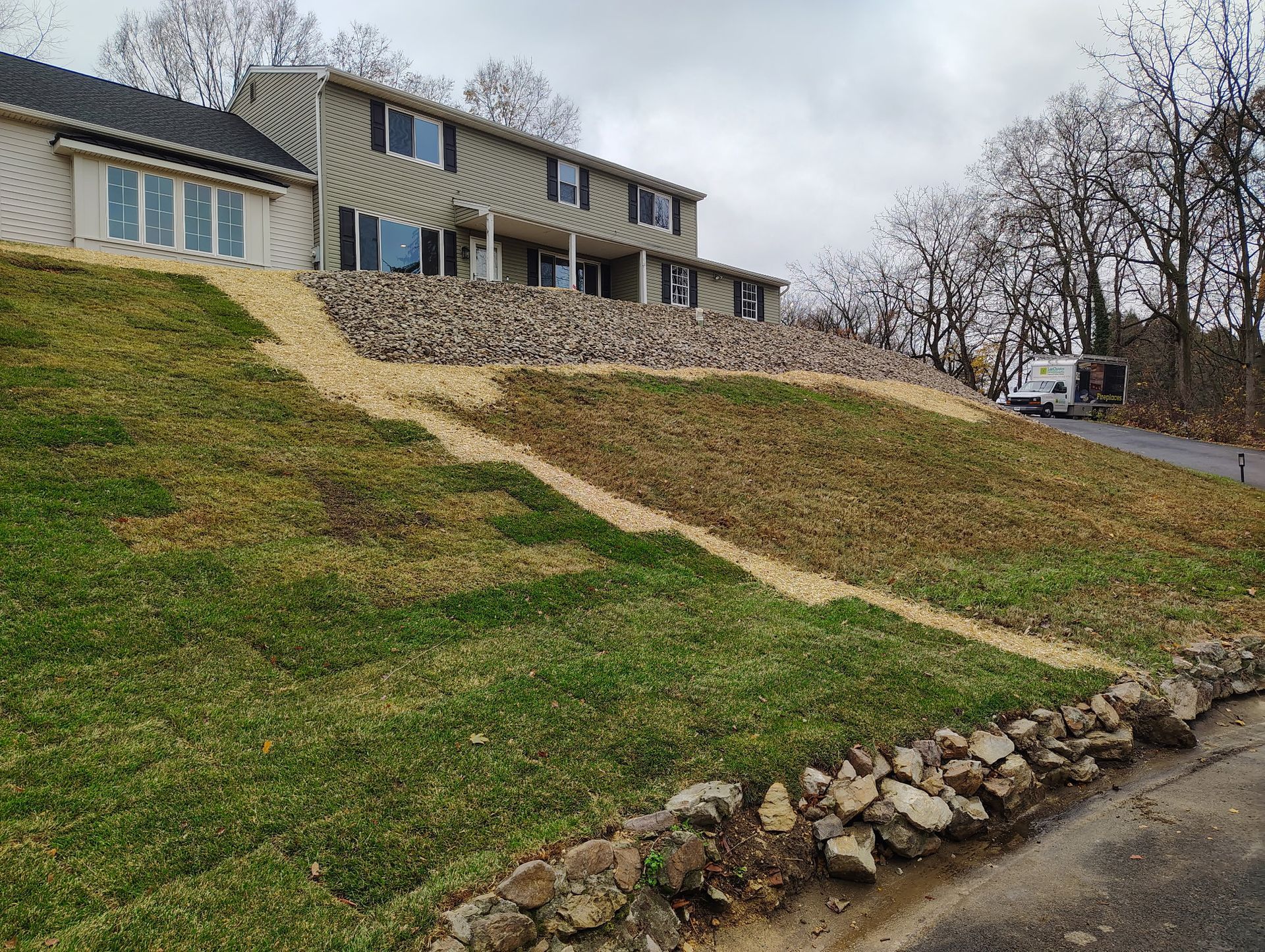 A tan house sits atop a grassy slope with a large rock retaining wall and a diagonal trench filled with sand or gravel.