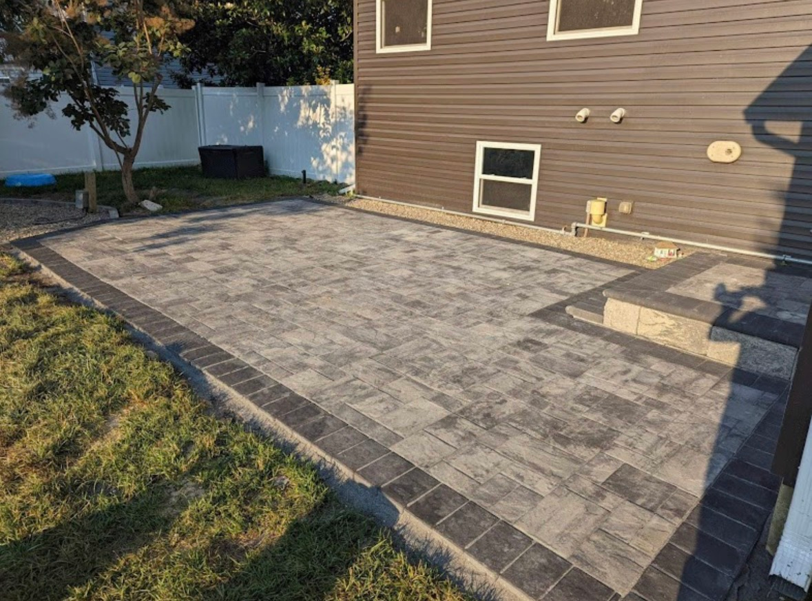 Paver patio beside a brown house, bordered by grass and a small concrete step area.