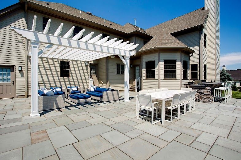Back patio with white pergola, blue lounge chairs, dining table, and stone pavers by a house