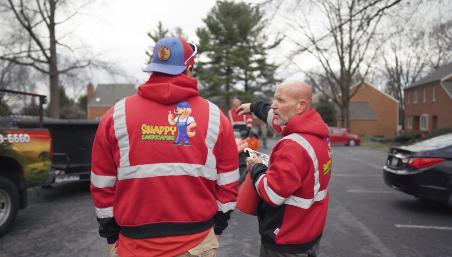 Two people in red jackets and beanies stand in a parking lot, one smiling and gesturing toward the other.