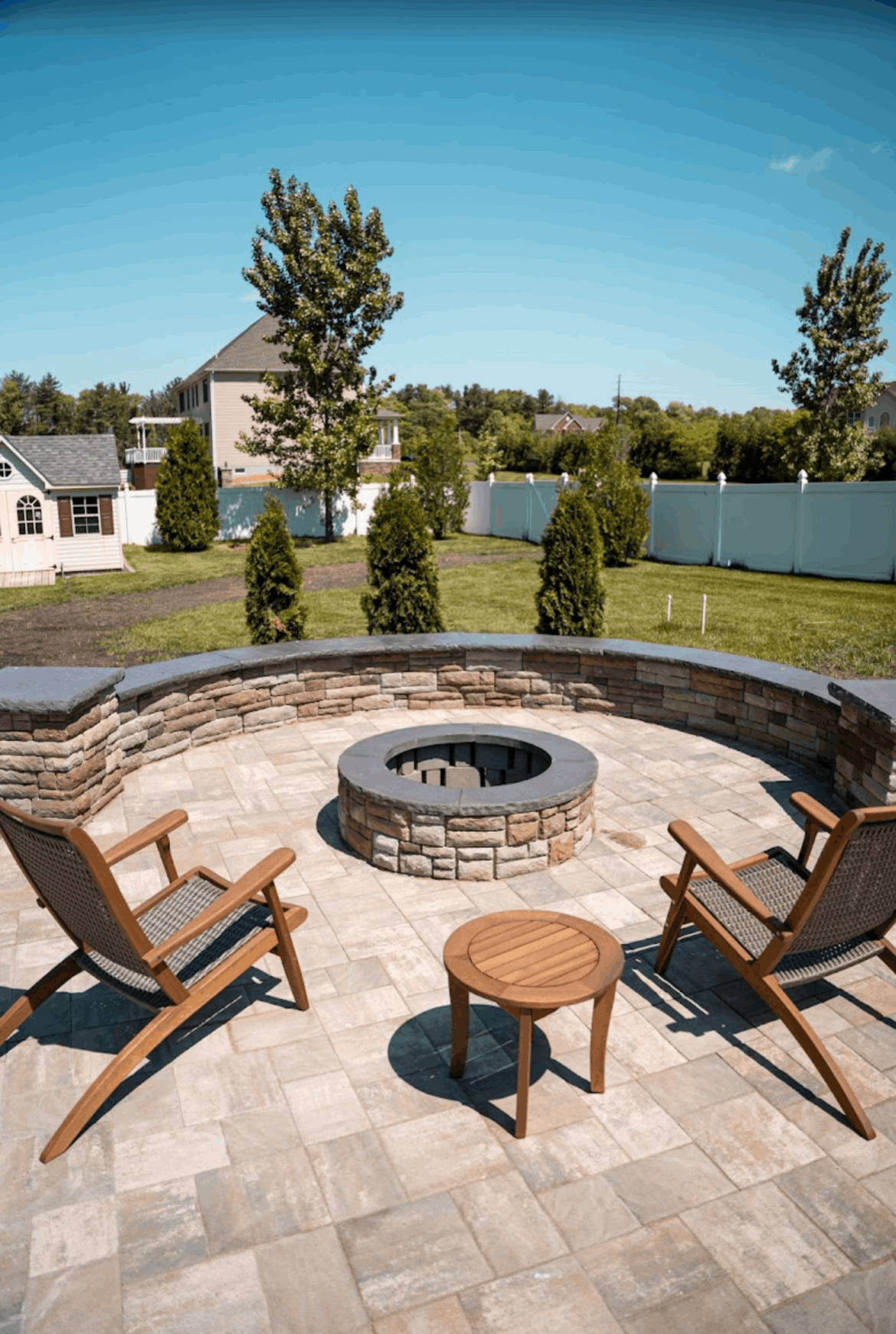 Stone fire pit with wooden chairs on a paved patio, overlooking a lake and grassy yard under a clear blue sky