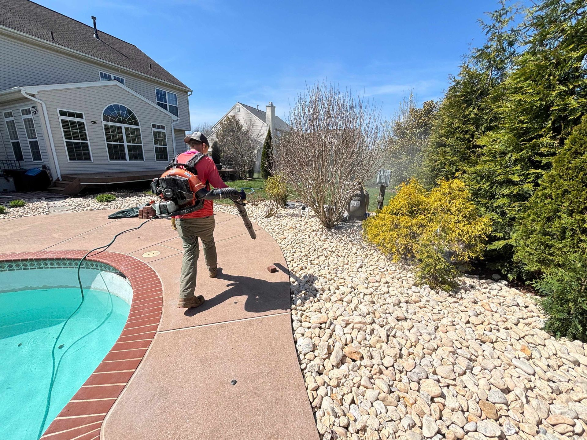 Person using a leaf blower beside a pool and stone landscaping on a sunny day