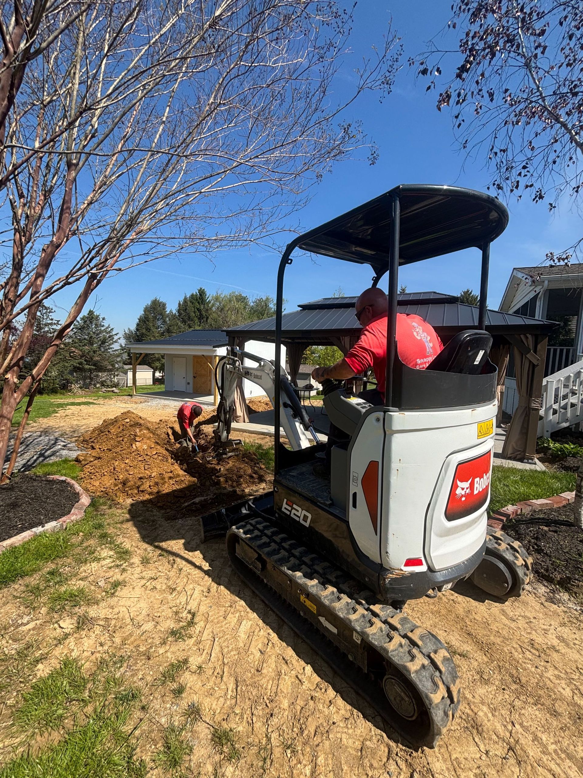 Mini excavator with a red bucket working in a sunny yard beside a tree and house.