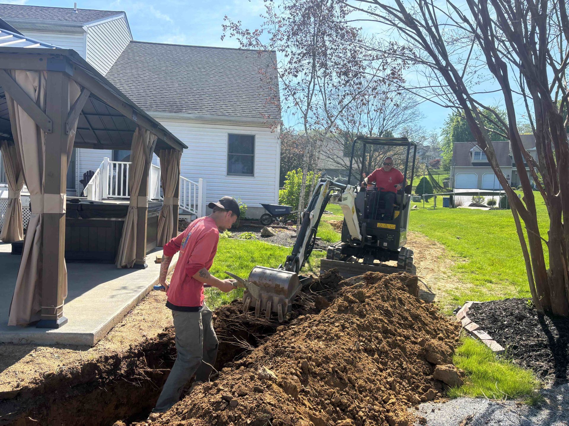 Two workers operating machinery to move a large dirt pile beside a house and deck