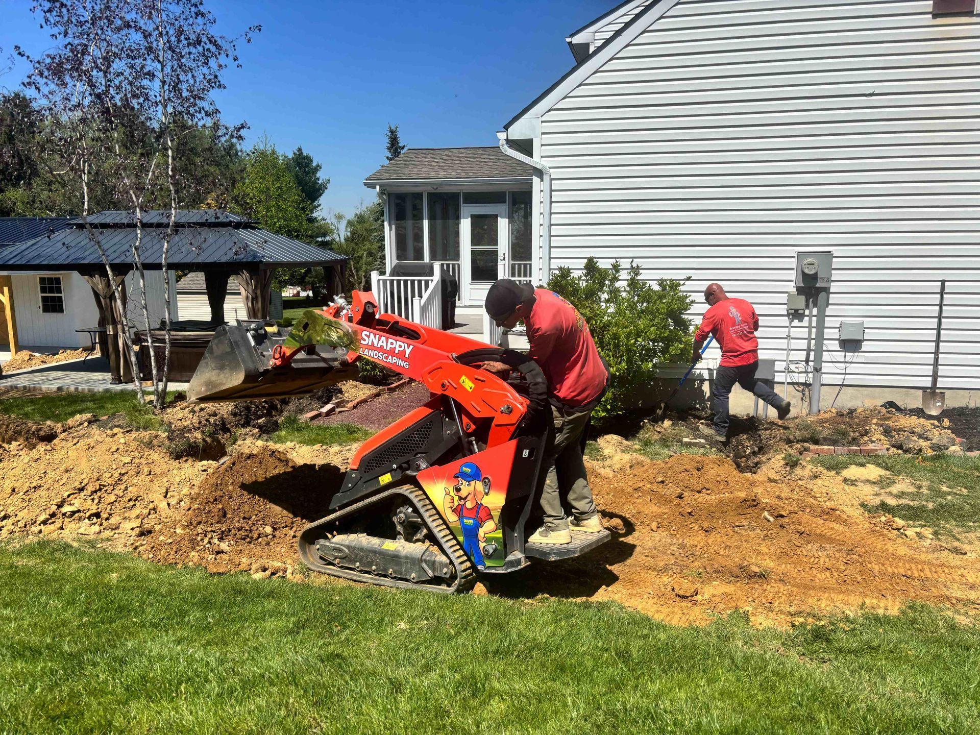 Workers operating a red mini excavator beside a house, moving dirt in a yard