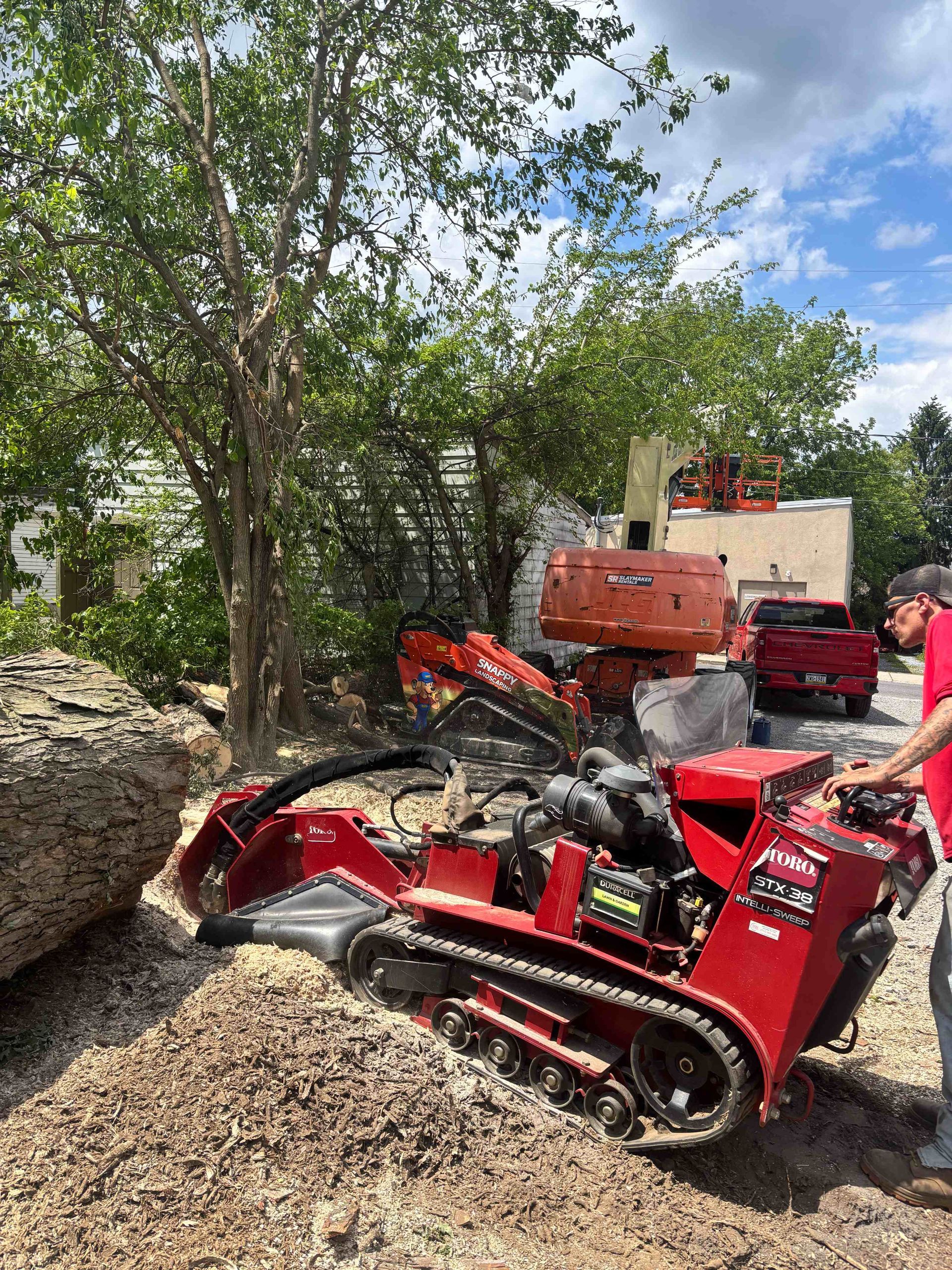 Red tracked stump grinder on gravel near trees, with orange and red equipment in the background.