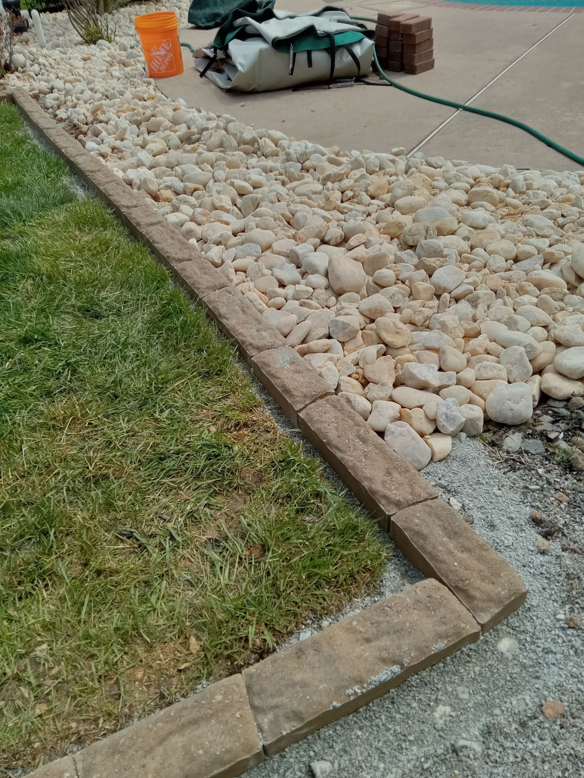 Landscaped walkway bordered by tan rocks, concrete edges, and green grass beside a driveway