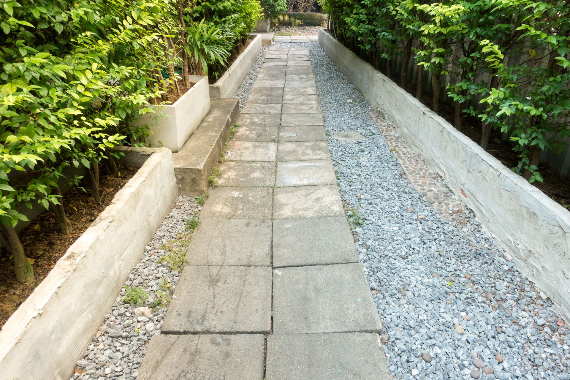 Narrow stone walkway bordered by lush green plants and a gravel path on the right