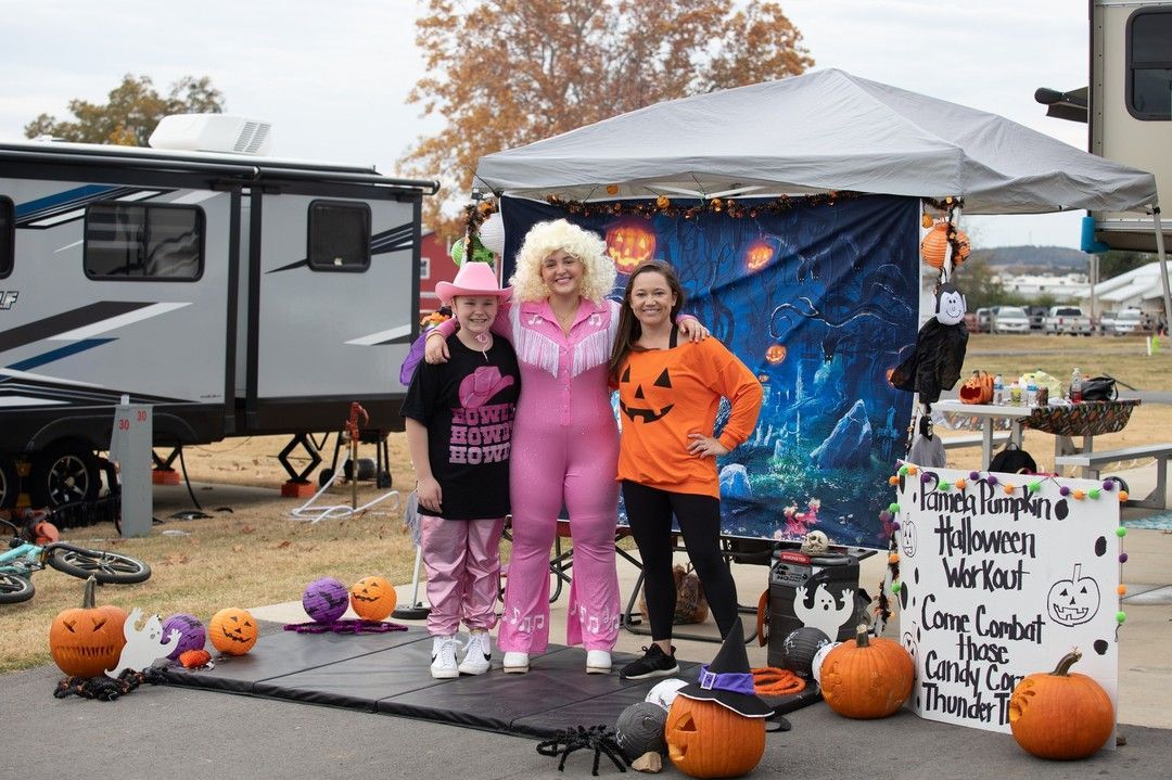 Three people pose at a Halloween campsite with pumpkins, a gray trailer, and a spooky backdrop.