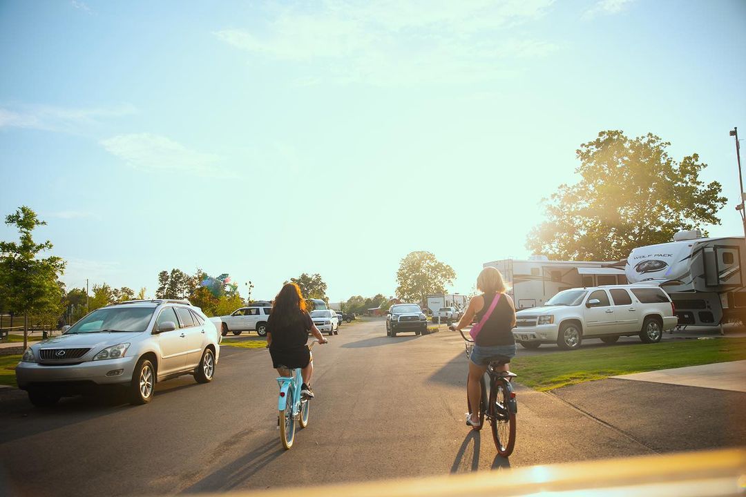Two cyclists ride down a sunlit street with parked cars and RVs at sunset.
