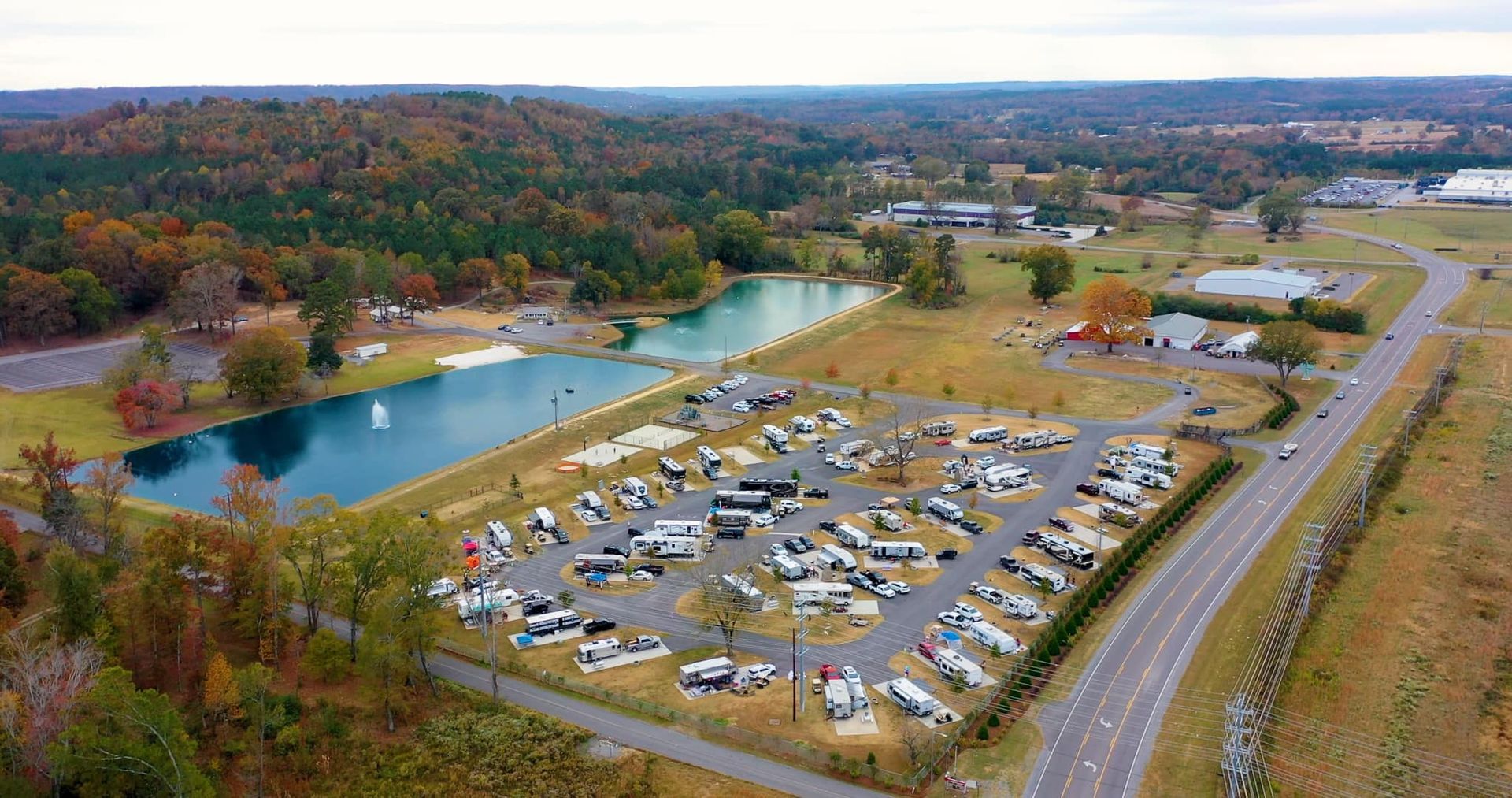 Aerial view of a lakeside campground with RVs, parked cars, and autumn trees beside a road