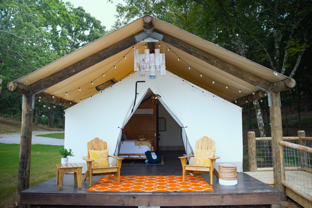 Open-front white glamping tent with two wicker chairs on a wooden deck in a wooded setting