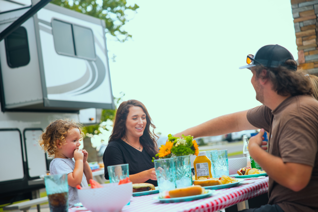 Family sharing a picnic at a campsite beside an RV, with drinks and snacks on a checkered tablecloth.