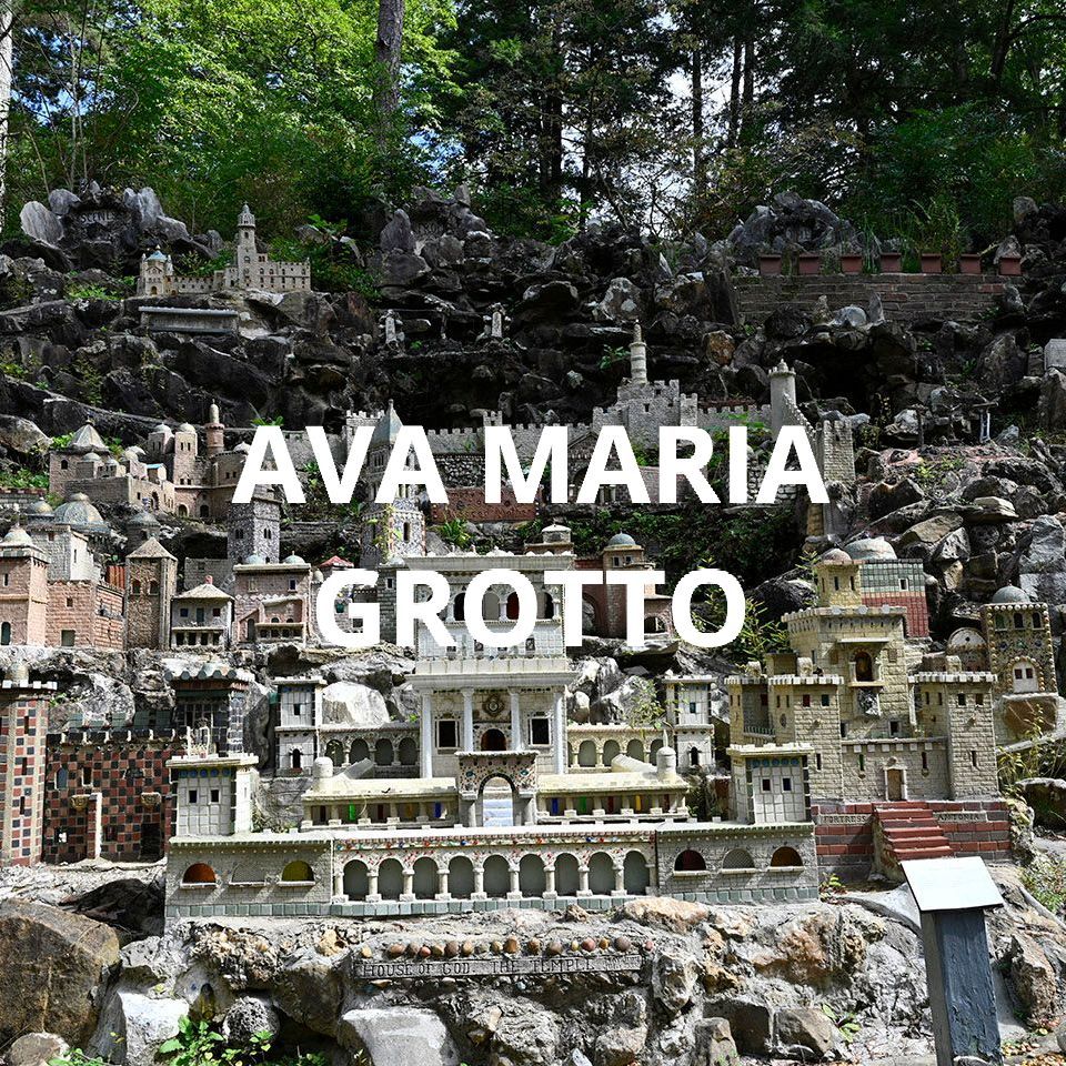 Miniature grotto display in a wooded garden, with large white text reading “AVA MARIA GROTTO.”