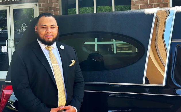 Man in a dark suit standing beside a black vehicle with an open door.