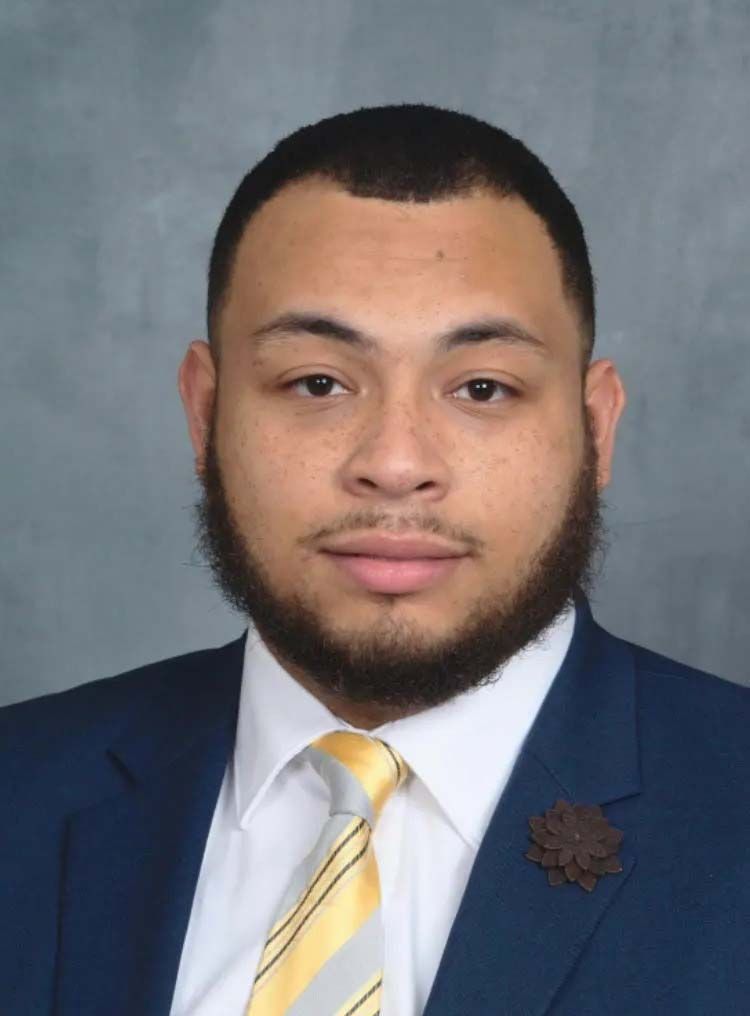 Studio portrait of a man in a navy suit, white shirt, and yellow striped tie, against a gray background