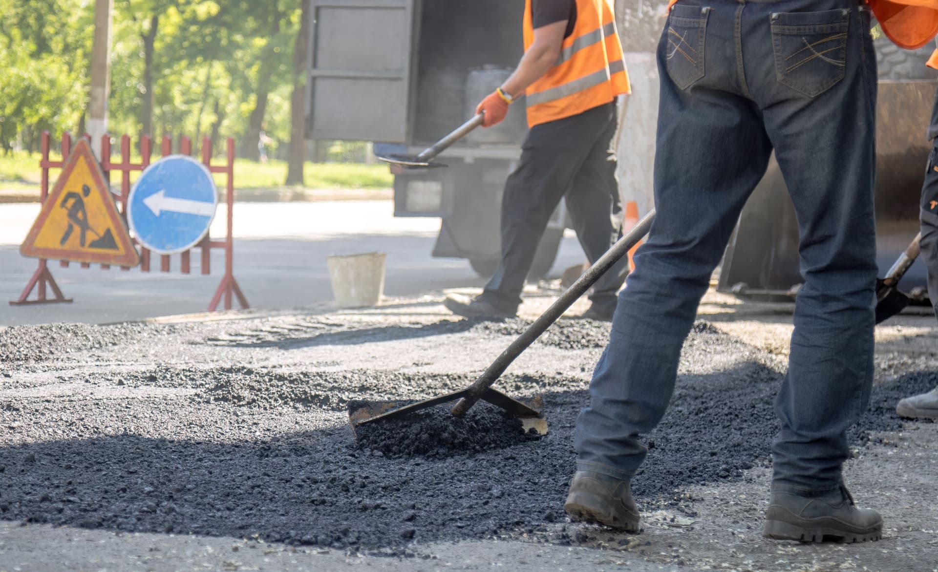 Road construction worker using a rake to level fresh asphalt with warning traffic signs in the background