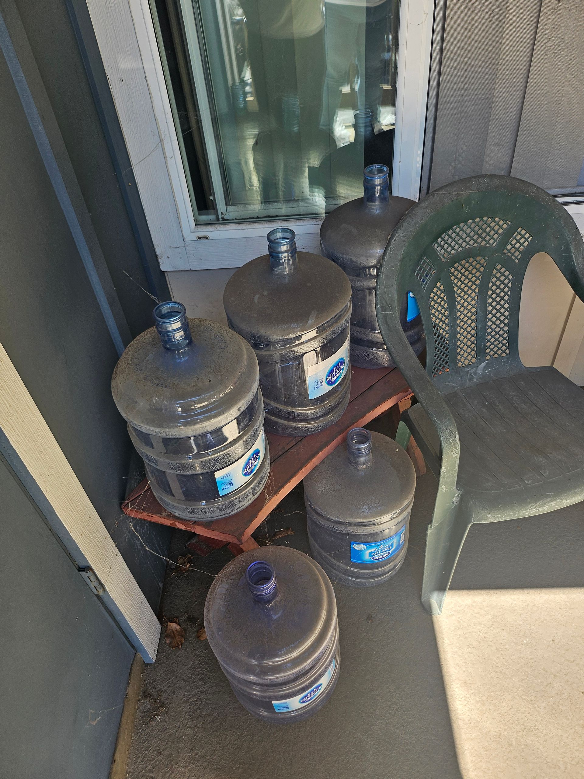 Several large water jugs on a balcony beside a green plastic chair and glass door