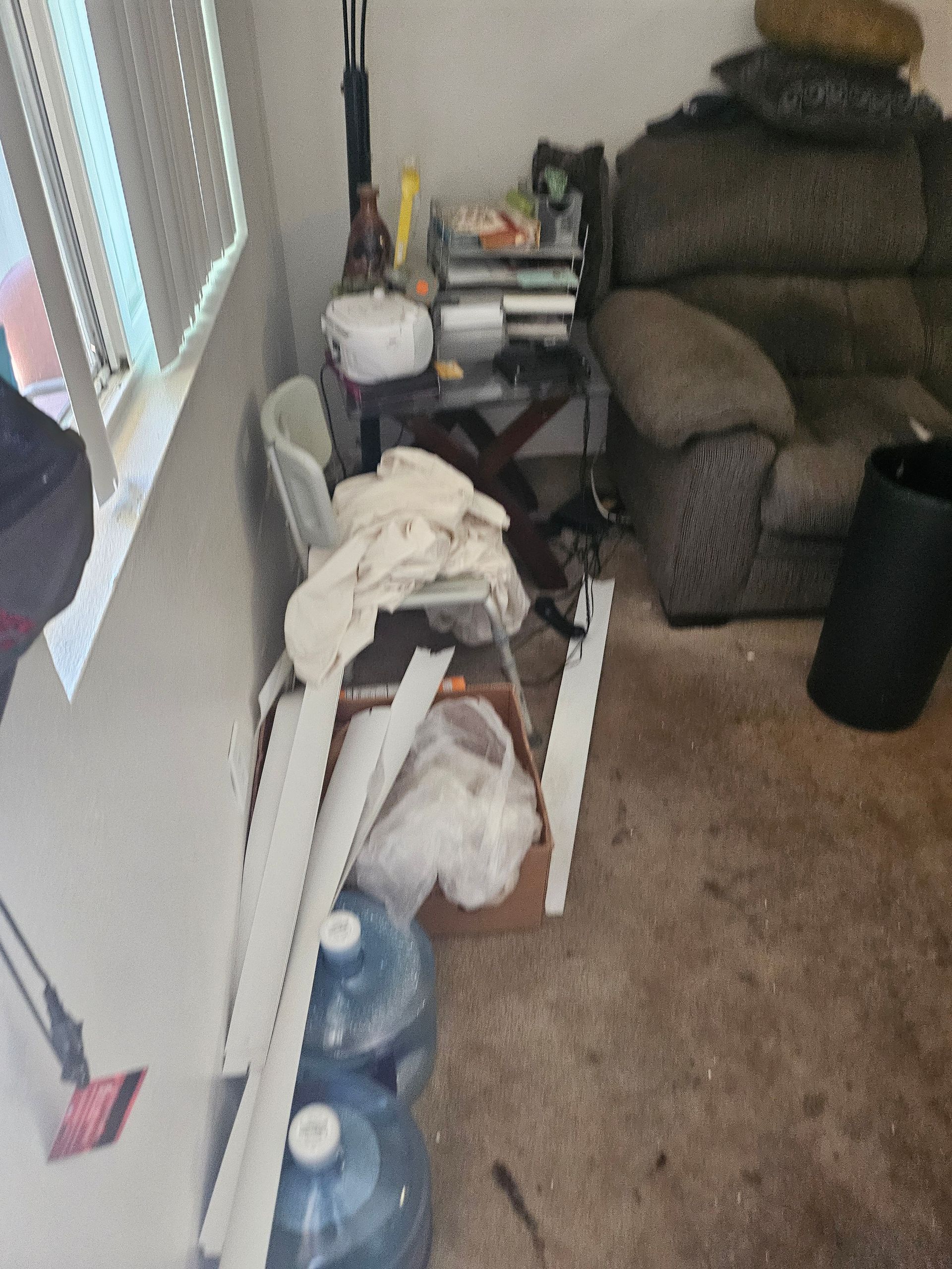 Cluttered living room corner with stacked items beside a recliner, window, and vacuum on a carpeted floor