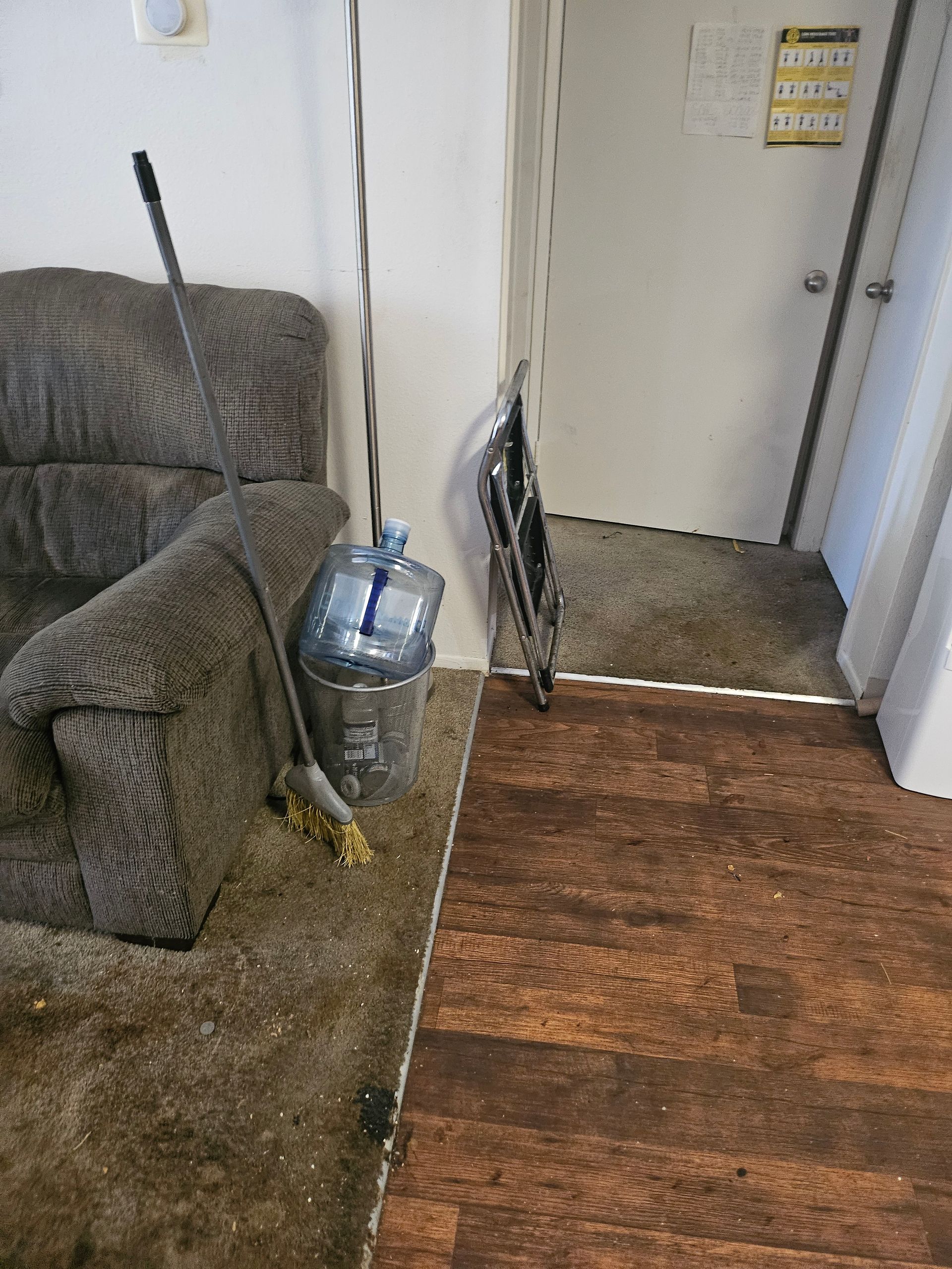 Cluttered living room corner with a gray recliner, metal container, broom, and leaning fan by a doorway.
