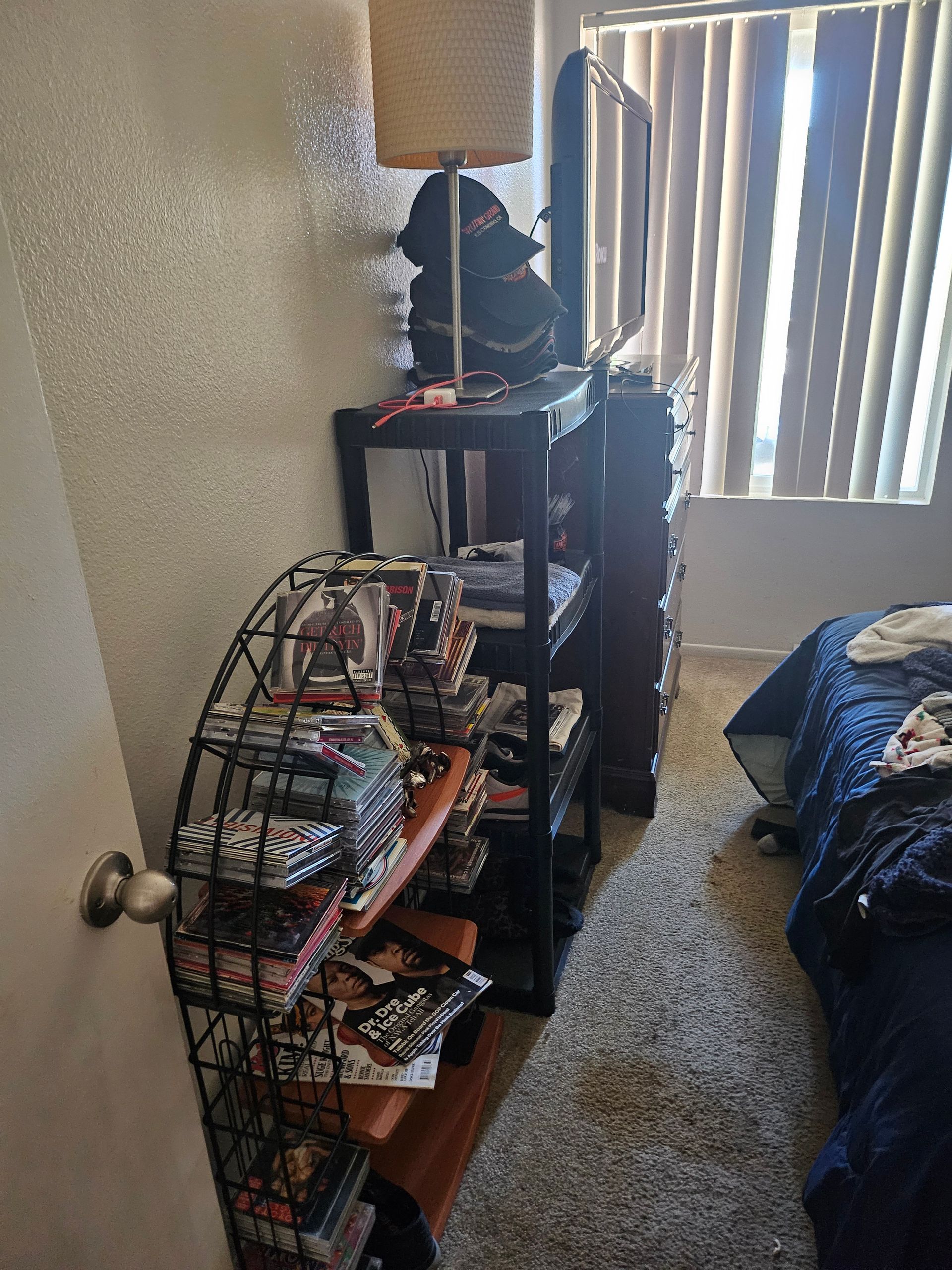 Small bedroom with a bunk bed, shelving unit, laundry rack, and a window with vertical blinds.