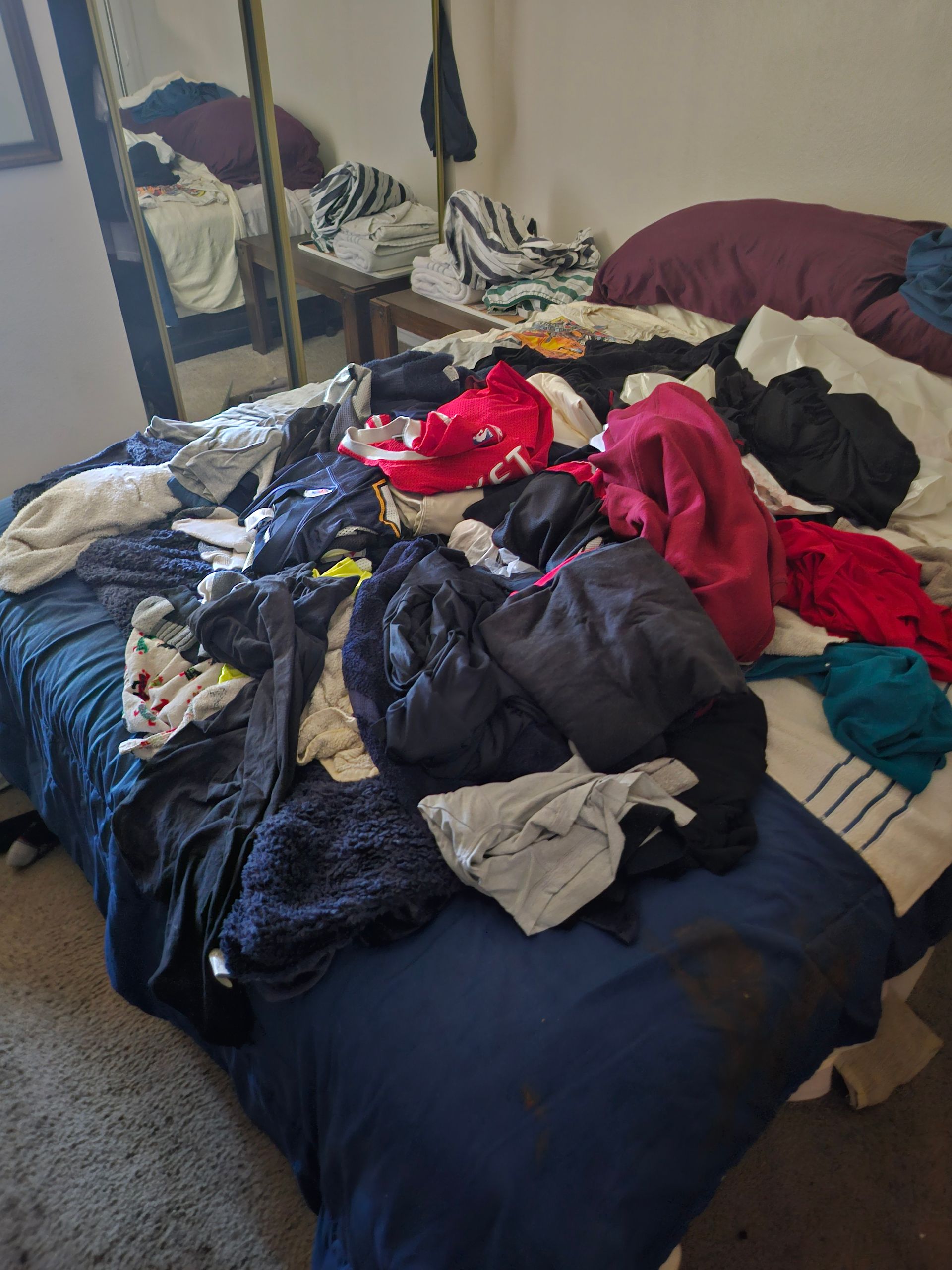 Bed with a large pile of colorful clothes and blankets scattered across it, beside a mirrored dresser.