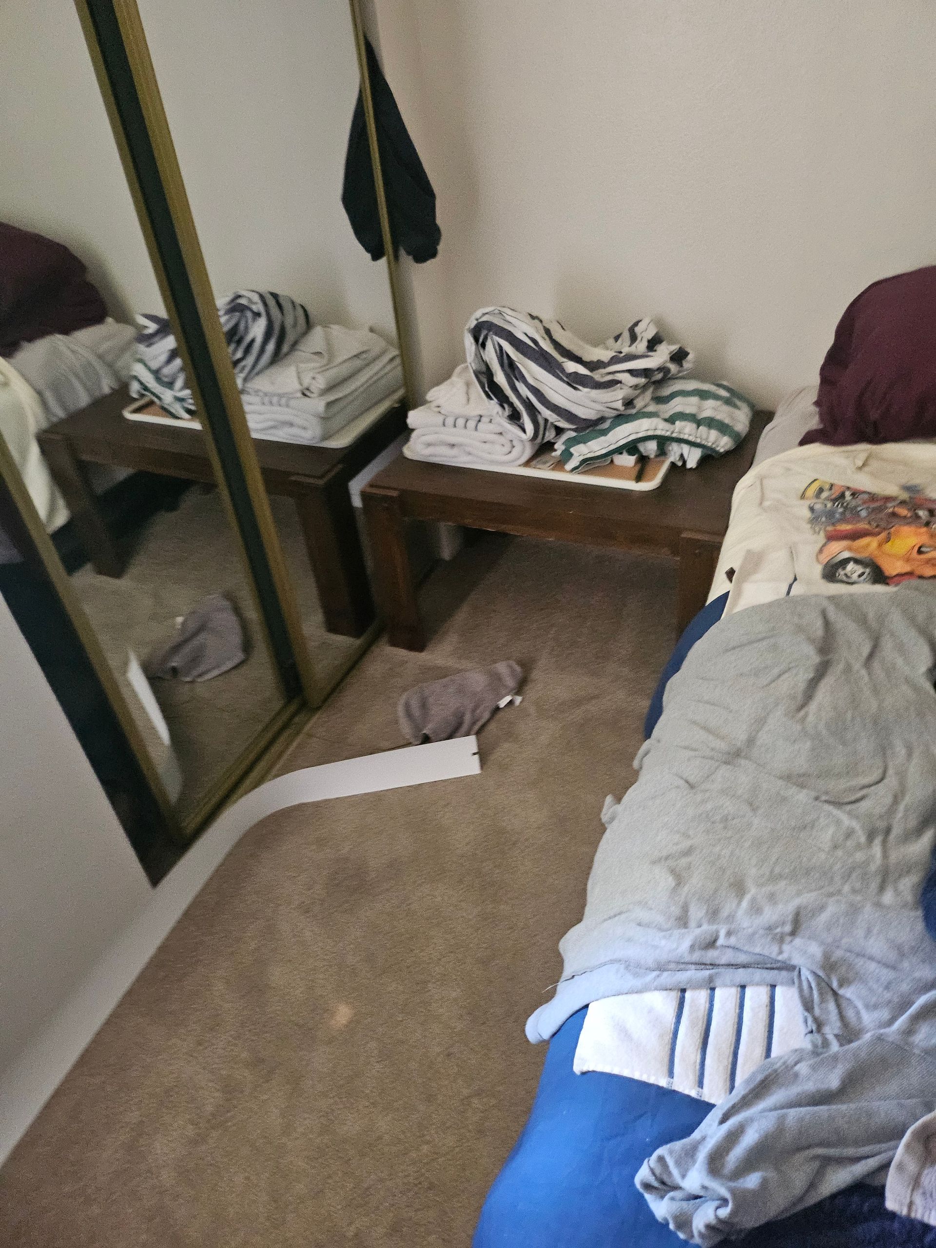 Small bedroom with an unmade bed, clothes piled on surfaces, and a mirror against the wall.
