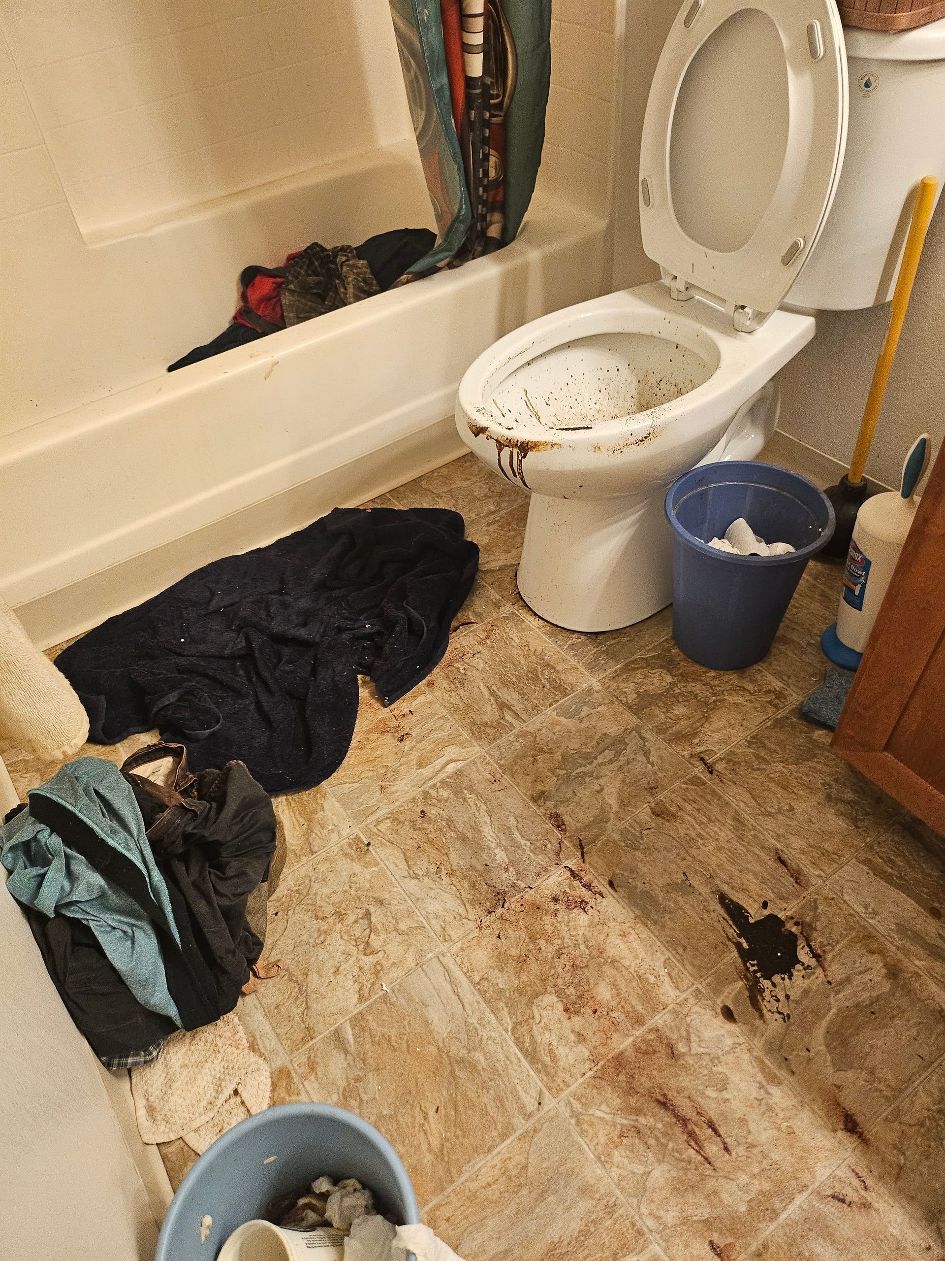 Messy bathroom with toilet, dirty floor, laundry, and a bucket beside the tub