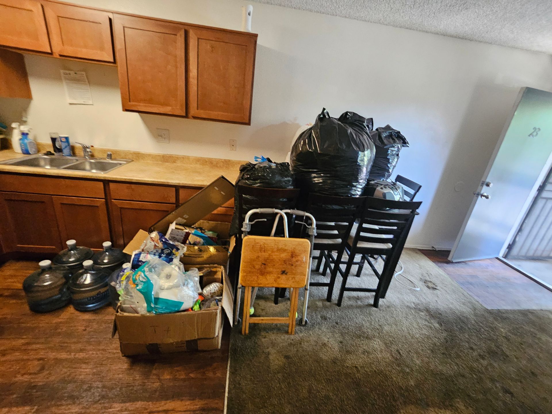 Cluttered kitchen with stacked boxes, bags, chairs, and pots on the floor near cabinets and counters