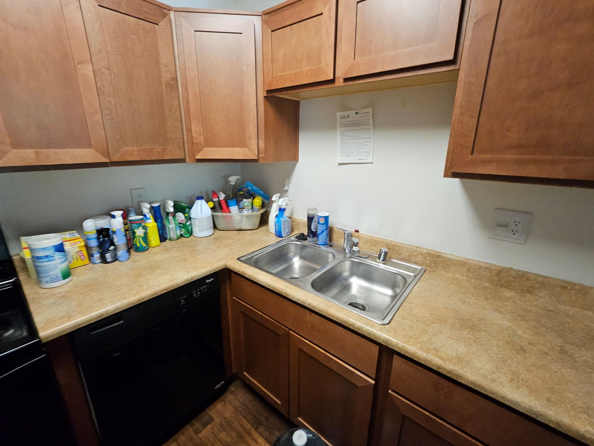 Small kitchen corner with wooden cabinets, tan counters, sink, and a row of cleaning supplies on the counter