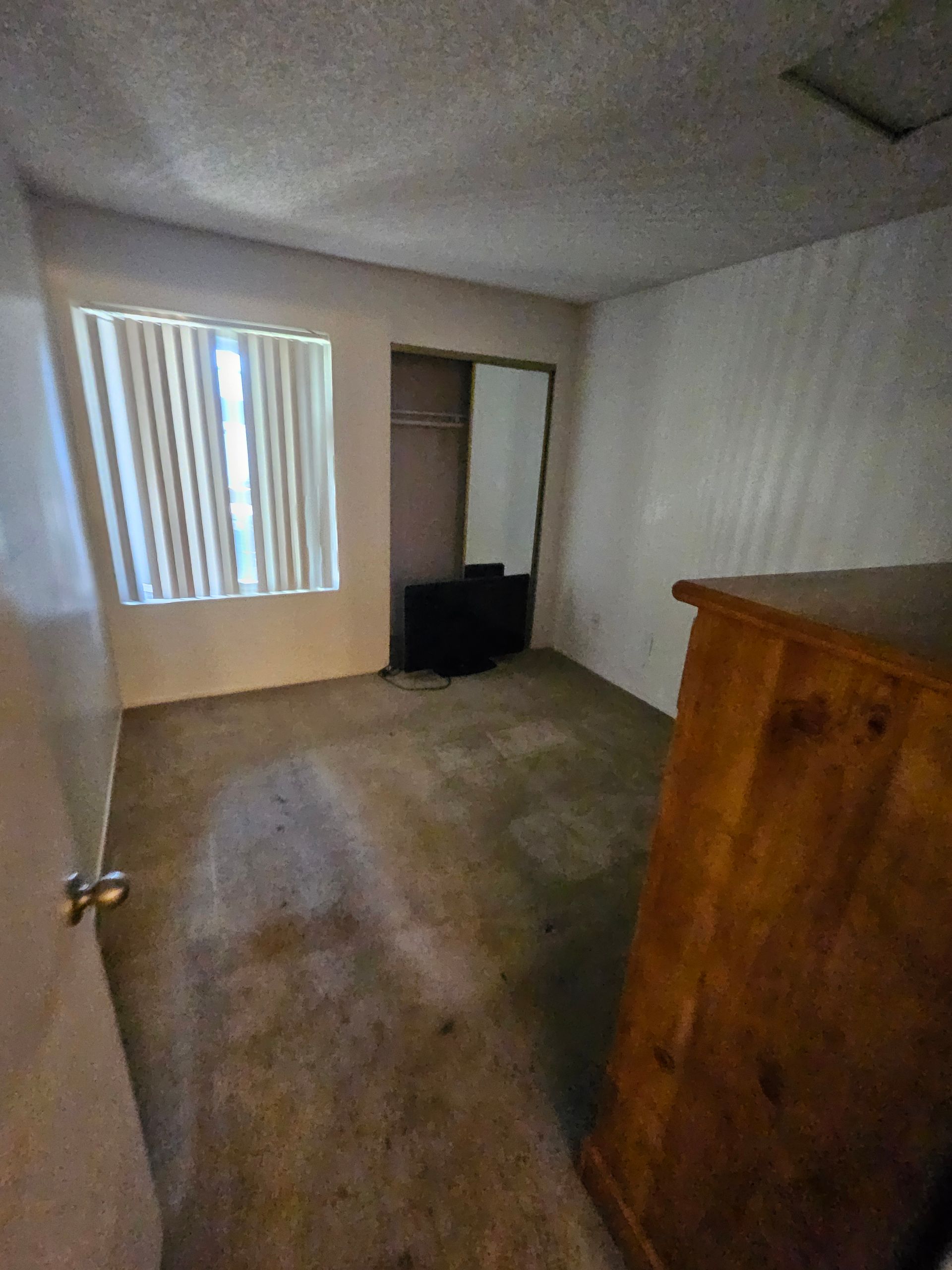 Empty carpeted bedroom with striped window blinds, closet opening, and wooden dresser on the right.