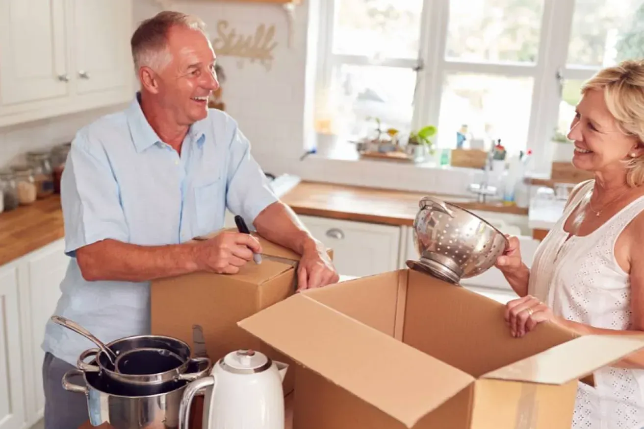 Two people unpacking boxes in a bright kitchen, smiling beside cookware and a window.