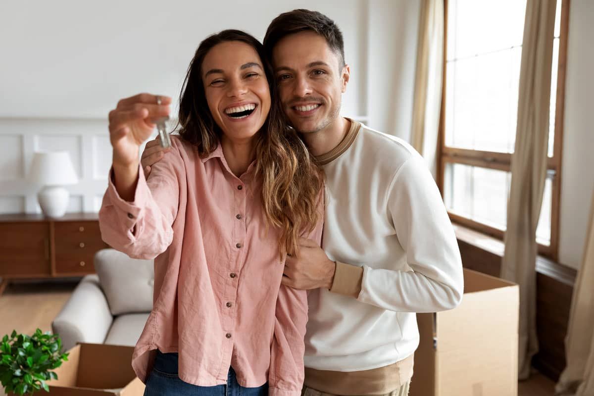 Smiling couple posing together in a bright living room, one in a pink shirt and one in a cream sweater.