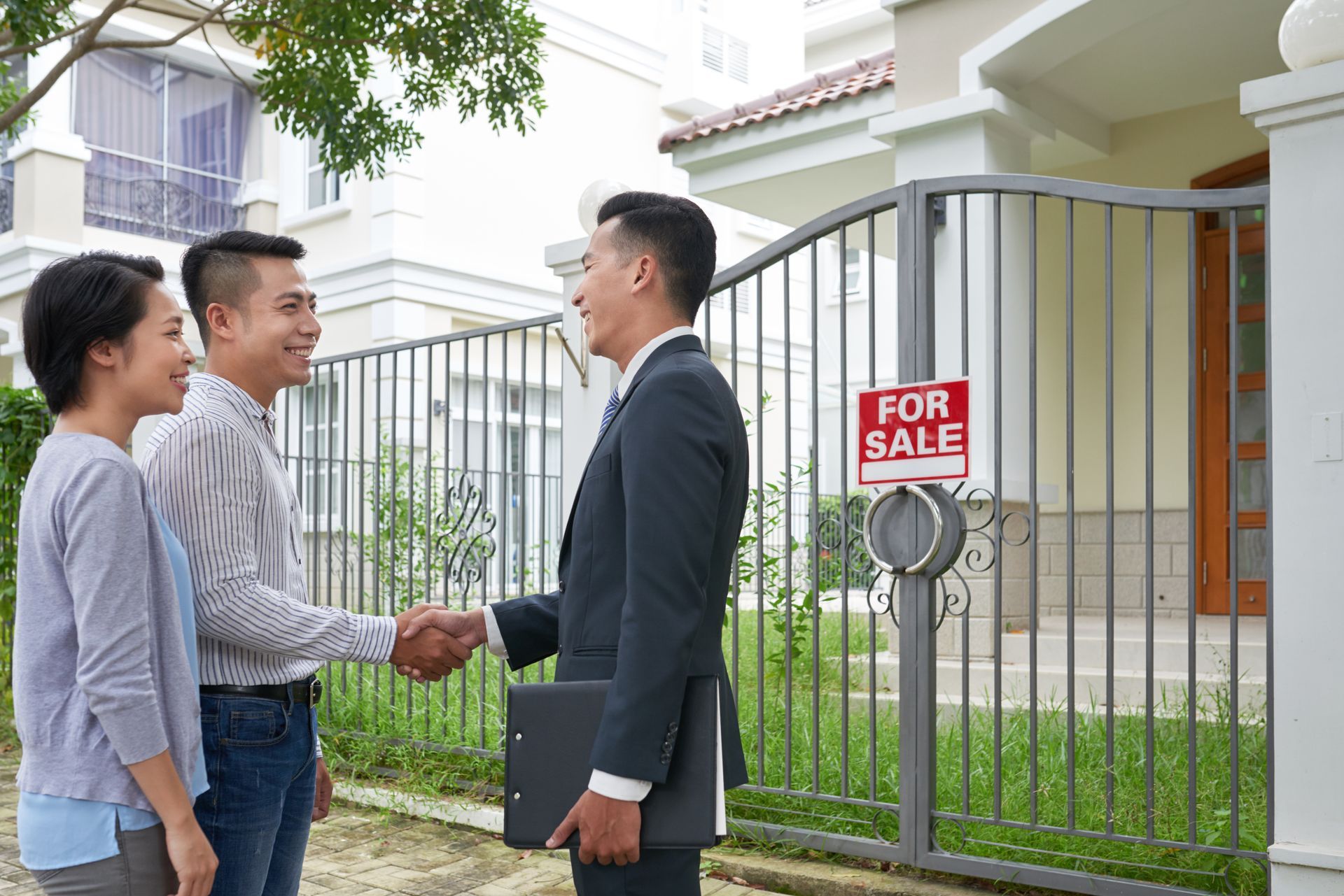 Two men shaking hands outside a house for sale while a woman stands nearby by the gate
