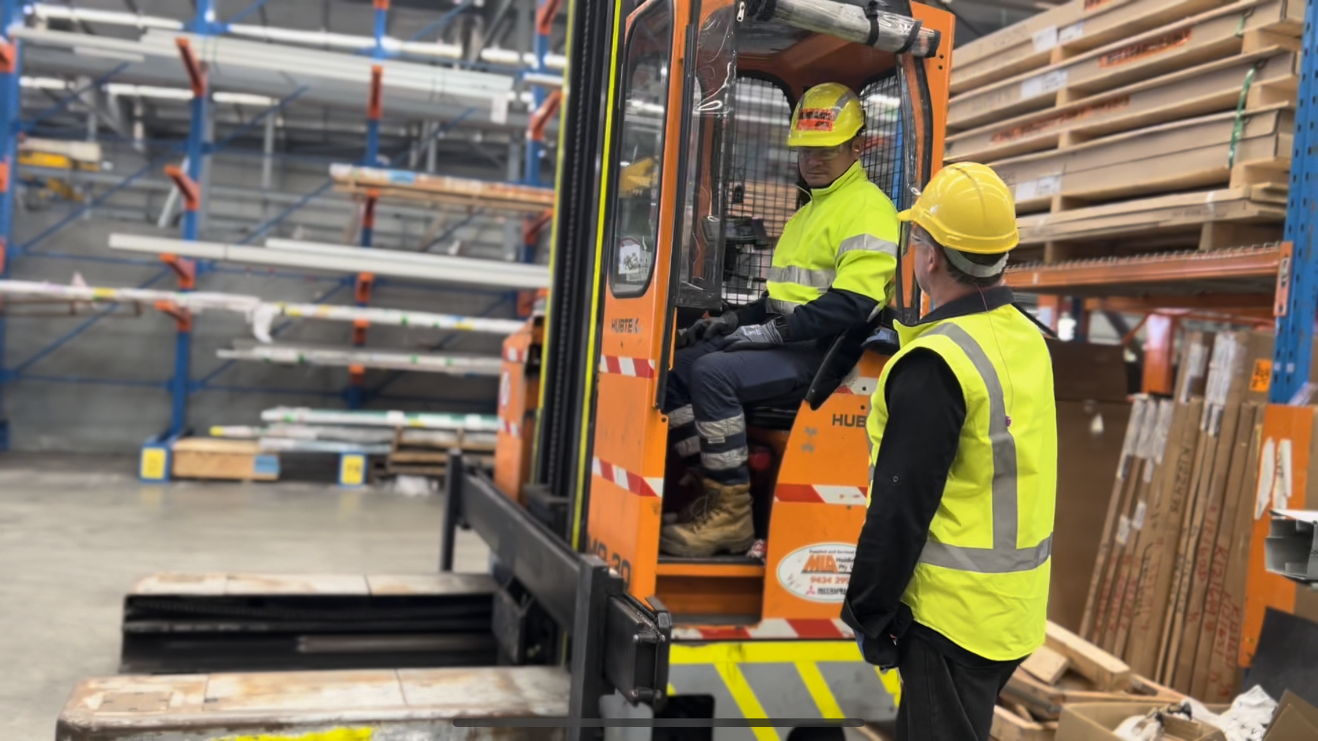 A man wearing yellow hi-vis and a yellow hard hat standing in a warehouse talking to another man in yellow hi-vis and a yellow hard hat who is sitting on an orange forklift