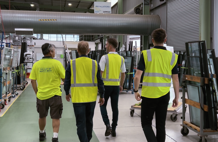a group of 4 men in a glass manufacturing warehouse wearing yellow hi vis and walking away from the camera