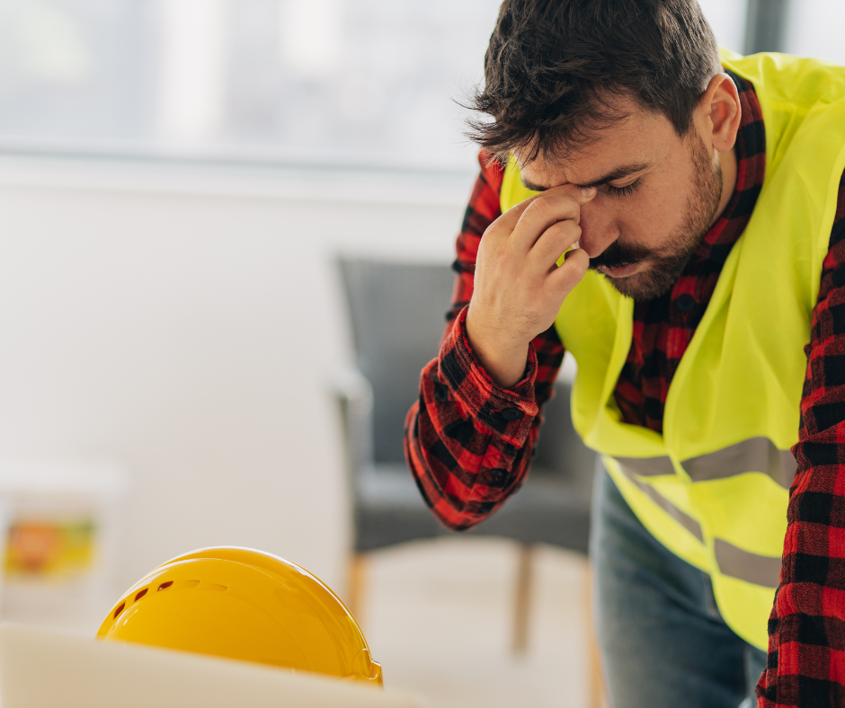 Electrical worker dressed in a red and black shirt with a yellow hi-vis with a yellow construction helmet beside him while looking stressed out