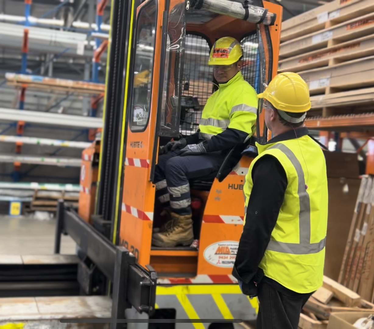 A man wearing yellow hi-vis and a yellow hard hat in a warehouse talking to another man wearing a yellow hi-vis jumper, yellow hard hat, navy work pants and safety boots driving an orange forklift
