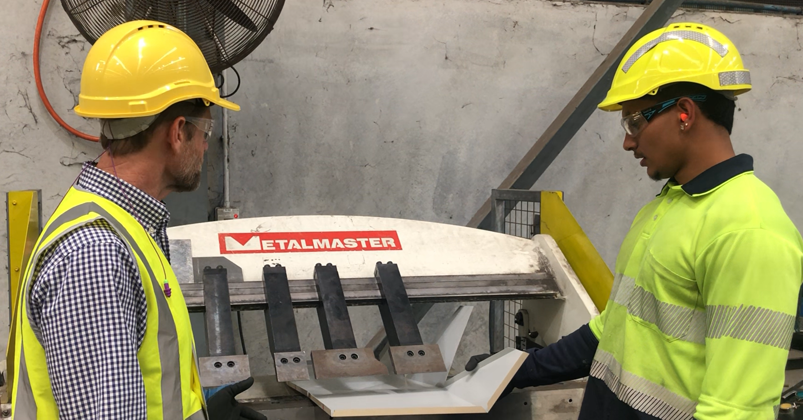 2 men wearing yellow hi-vis, hard hats and safety glasses looking at a sheetmetal bending machine in a manufacturing warehouse